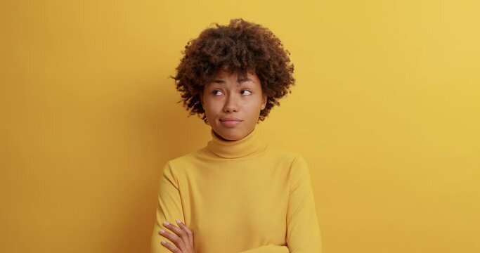 Hesitant uncertain woman looks clueless at camera says I dont care keeps arms folded and gazes puzzled at camera isolated over yellow background. Slow motion. Female shrugs shoulders in bewilderment