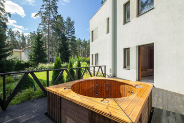 Modern exterior of luxury private house. Wooden bath tub on terrace. Huge garden with green grass and trees.
