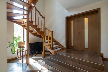 Luxury interior of entrance hall in apartment. Wooden staircase. Huge door. Green plant in pot.