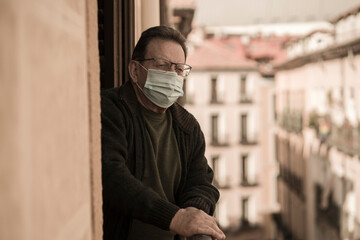 mature man in balcony home lockdown during covid19 outbreak - senior male on his 70s in face mask worried and tired looking to the street thoughtful and depressed in quarantine