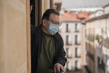mature man in balcony home lockdown during covid19 outbreak - senior male on his 70s in face mask worried and tired looking to the street thoughtful and depressed in quarantine