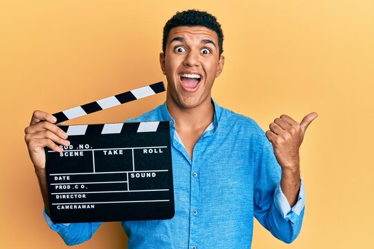 Young arab man holding video film clapboard pointing thumb up to the side smiling happy with open mouth