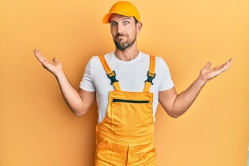 Young handsome man wearing handyman uniform over yellow background clueless and confused expression with arms and hands raised. doubt concept.