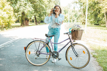 A stylish woman standing near a bicycle while using a retro camera.