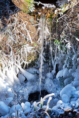 Water flow falls down from old rusty pipe into small puddle covered with white ice and hoarfrost on dry grass in cold winter forest at sunlight extreme closeup.