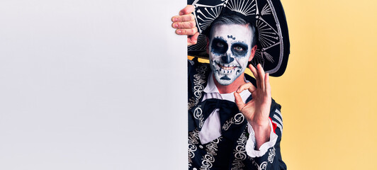 Young man wearing mexican day of the dead costume holding blank empty banner doing ok sign with fingers, smiling friendly gesturing excellent symbol