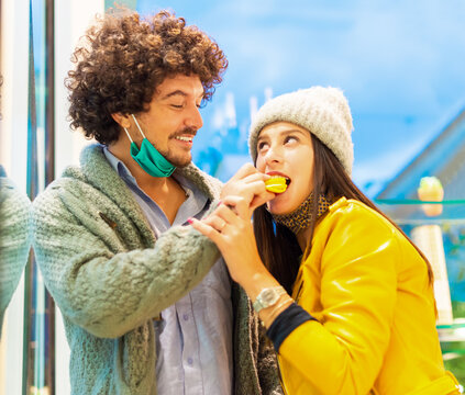 Young and happy couple wearing protective face mask eating a macarones standing inside a shop.girl having a sweet  dessert candy in autumn after lockdown reopening. concept about joy and happiness - Powered by Adobe