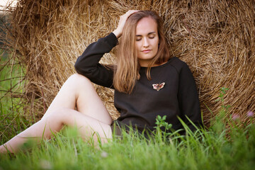 woman in a black sweater with a handmade brooch sits the the field. Young beautiful brunette woman posing on a background of haystacks in a cut field