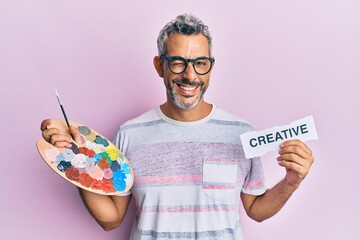 Middle age grey-haired man holding palette and creative message paper winking looking at the camera...