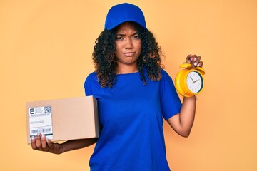 Young african american woman holding delivery package and alarm clock skeptic and nervous, frowning...