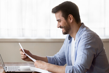 Fototapeta na wymiar Great news. Happy satisfied millennial businessman remote worker reading paper postal letter message notification containing information he is hired promoted receives financial reward high dividends