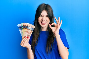 Young beautiful caucasian girl holding canadian dollars doing ok sign with fingers, smiling friendly gesturing excellent symbol