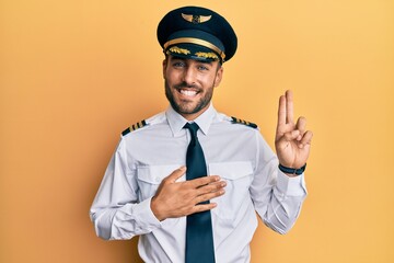 Handsome hispanic man wearing airplane pilot uniform smiling swearing with hand on chest and...