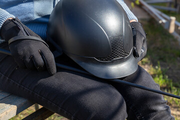 Woman in black gloves sitting on the wooden bench and holding helmet and whip in her hands....