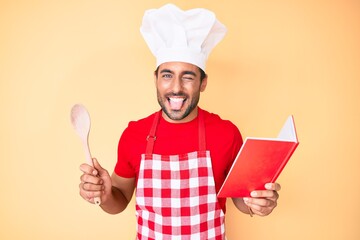 Young hispanic man wearing professional baker apron reading cooking recipe book sticking tongue out happy with funny expression.