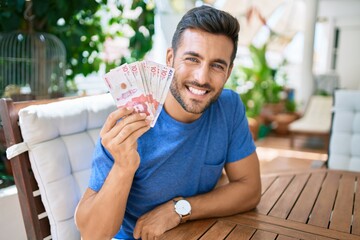 Young hispanic man smiling happy holding colombian pesos banknotes at the terrace.