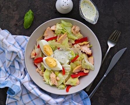 Healthy salad with iceberg lettuce, boiled salmon, sweet pepper and egg in a gray plate on a dark concrete background. Salad recipes.