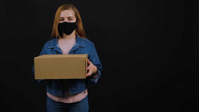 Woman wearing face mask while holding a package and standing in the studio. Isolated on black background