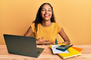 Young african american girl working at the office with laptop and calculator smiling and laughing hard out loud because funny crazy joke with hands on body.