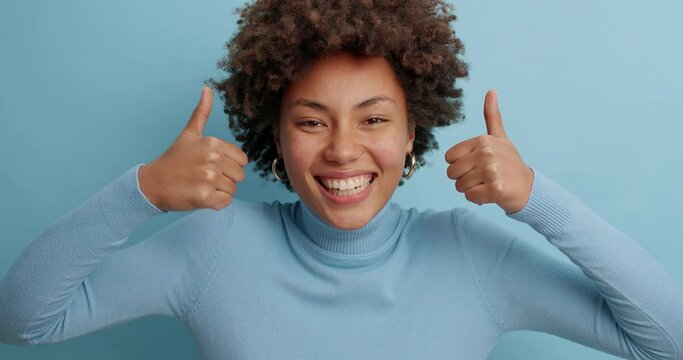 Optimistic dark skinned woman makes thumbs up gives approval shows excellent gesture says nice job gives positive feedback shares good opinion dressed in casual wear isolated over blue background
