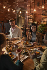 Vertical portrait of cheerful adult people sitting at dinner table while enjoying party with outdoor lighting