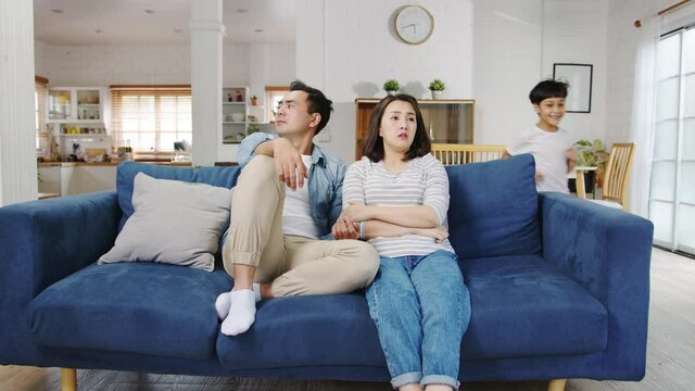 Asia family dad and mom sit on couch feels annoyed exhausted while daughter and son have fun shouting run around sofa in living room at home. Social distancing, Quarantine for corona virus prevention.
