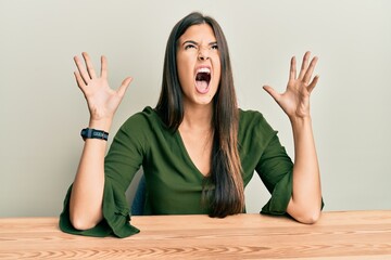 Young brunette woman wearing casual clothes sitting on the table crazy and mad shouting and yelling...