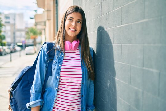Young hispanic student girl smiling happy using headphones leaning on the wall at the city.