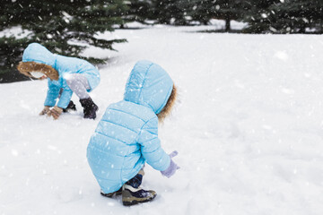 Fototapeta na wymiar Little girls, sisters are making snowman in snowy winter park. walking, Having fun. Stylish clothes, jackets with fur, pants with snowflakes, mittens. Family picnic in cold weather. Outdoor activity