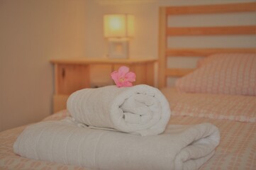 Fototapeta na wymiar Rolled white towel on single wooden bed with illuminated lamp in background. Detail of a modern double bedroom