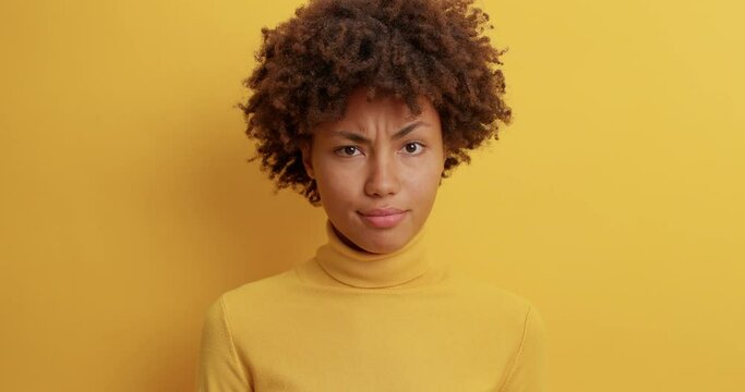 Dissatisfied dark skinned woman with Afro hair smirks face and says no looks displeased cant believe in situation mistrusts person telling lies wears casual turtleneck isolated on yellow background