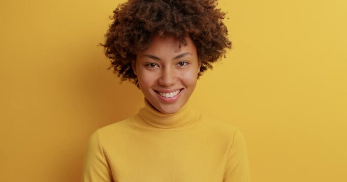 Lovely calm pleased woman with curly hair smiles broadly looks satisfied at camera being in good mood dressed in casual turtleneck expreses positive emotions isolated over yellow background.