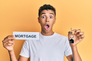 Young handsome african american man holding paper with mortgage word and house keys afraid and shocked with surprise and amazed expression, fear and excited face.