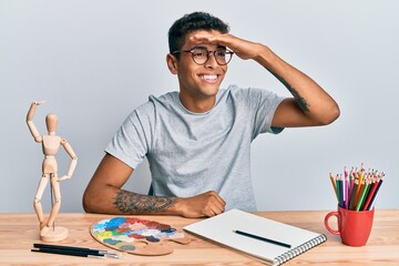 Young handsome african american man painter sitting palette and art manikin very happy and smiling looking far away with hand over head. searching concept.