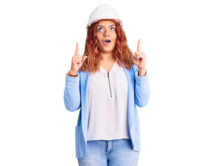 Young latin woman wearing architect hardhat amazed and surprised looking up and pointing with fingers and raised arms.