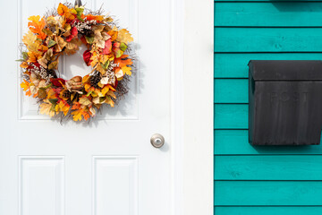 A wooden teal green exterior wall of a building with a white metal door. There's a wreath with orange autumn flowers hanging on the door. A black metal mailbox hangs to the right of the entrance. 