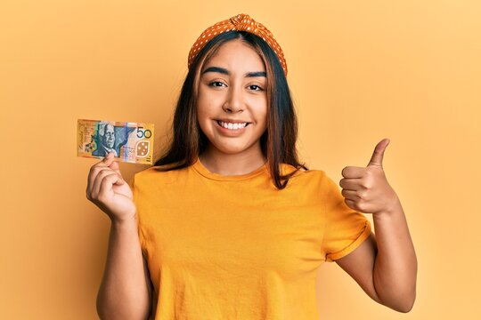 Young latin woman holding 50 australian dollar banknote smiling happy and positive, thumb up doing excellent and approval sign