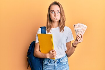 Beautiful blonde woman wearing student backpack and holding 50 turkish lira puffing cheeks with...