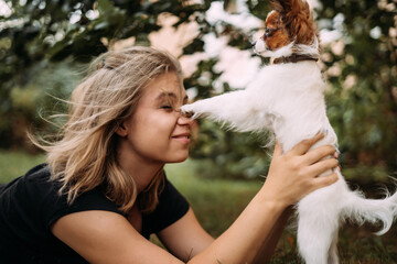 A woman lies on the grass holding a small puppy a white and red brown papillon playing. The dog is a companion friend of man.