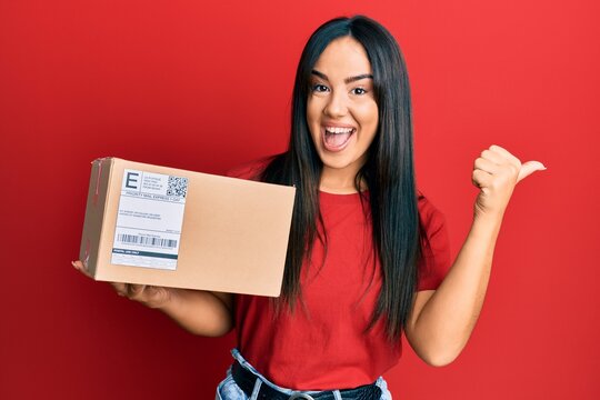 Young beautiful hispanic girl holding delivery package pointing thumb up to the side smiling happy with open mouth