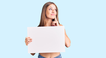 Young beautiful blonde woman holding blank empty banner serious face thinking about question with hand on chin, thoughtful about confusing idea
