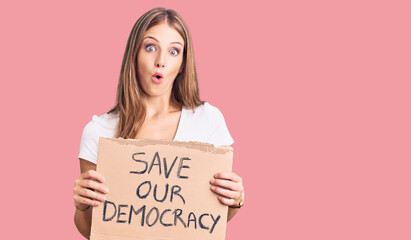 Young beautiful blonde woman holding save our democracy protest banner scared and amazed with open...