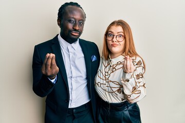 Young interracial couple wearing business and elegant clothes doing money gesture with hands, asking for salary payment, millionaire business