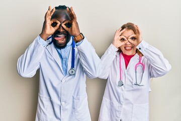 Young interracial couple wearing doctor uniform and stethoscope doing ok gesture like binoculars sticking tongue out, eyes looking through fingers. crazy expression.