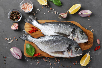 Fresh ready to cook raw fish dorado with ingredients and seasonings like basil, lemon, salt, pepper, red onion and garlic on wooden board on dark background, top view