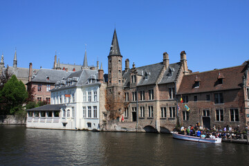 Naklejka na ściany i meble River side view of the old historic buildings of Bruges in Belgium on a sunny day. There is a boat with tourists on the river.