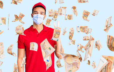 Young handsome hispanic man wearing delivery uniform and medical mask smiling with happy face winking at the camera doing victory sign. number two.