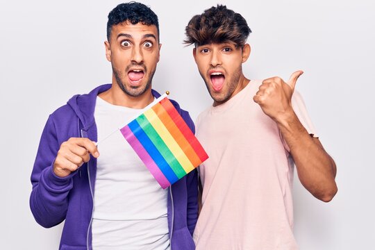 Young gay couple holding rainbow lgbtq flag pointing thumb up to the side smiling happy with open mouth