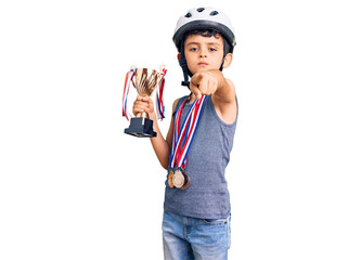 Little cute boy kid wearing bike helmet and winner medals holding winner trophy pointing with finger to the camera and to you, confident gesture looking serious