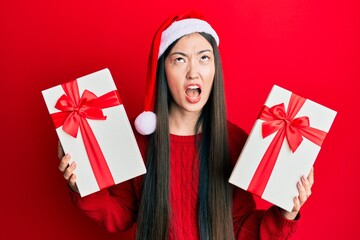 Young chinese woman wearing christmas hat and holding gifts angry and mad screaming frustrated and furious, shouting with anger looking up.
